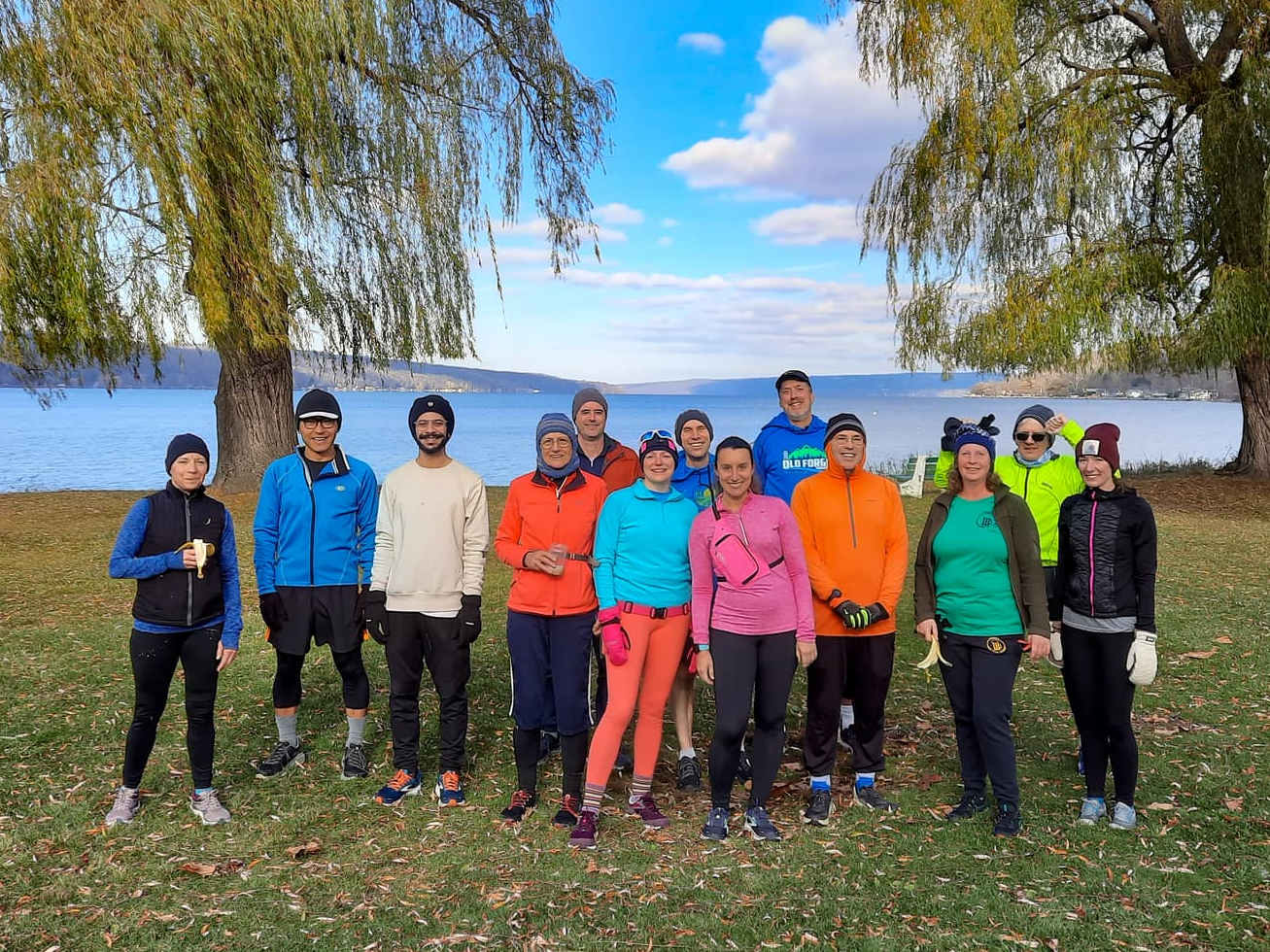 A group of smiling runners gathered together for a picture. Cayuga Lake is behind them.